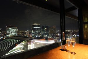 梅田駅1分、クラフトビールが自慢のお店『molto!!梅田店』にて、ディナー時に見られる大阪の夜景の画像をバックに、ワインボトルと2つのワイングラスを並べている画像
